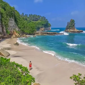 Pantai Ujung Genteng dengan hamparan pasir putih yang luas, air laut yang jernih, dan formasi batuan unik di kejauhan.
