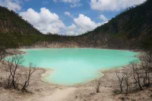 Pemandangan Kawah Putih di Ciwidey dengan danau berwarna turquoise yang dikelilingi tebing curam dan pepohonan gundul, di bawah langit biru berawan.
