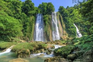 Tiga aliran air terjun Curug Cikaso yang megah jatuh ke kolam alami berwarna biru kehijauan, dikelilingi oleh vegetasi hijau.