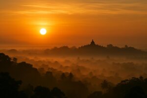 Pemandangan matahari terbit dari Punthuk Setumbu dengan siluet Candi Borobudur di kejauhan, kabut pagi, langit oranye keemasan.